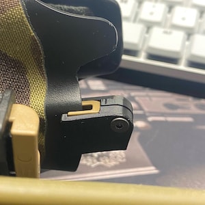 May include: Close-up of a black and tan tactical belt buckle. The buckle features a tan-colored release mechanism and a black body. The belt material has a camouflage pattern. A white keyboard is visible in the background.