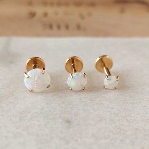 May include: Three gold stud earrings, each featuring a round, iridescent white opal. The earrings are presented on a light surface, highlighting their delicate design and subtle sparkle. The studs vary slightly in size.