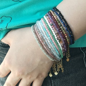 May include: A stack of colorful beaded bracelets with gold heart charms. The bracelets are made of various gemstones, including amethyst, rose quartz, and turquoise.