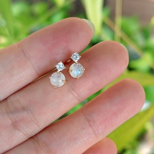 May include: A pair of gold-coloured stud earrings. Each earring features a round, iridescent moonstone and a small, square-cut diamond. The earrings are set against a blurred green background.