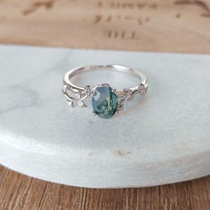 May include: A silver ring with an oval moss agate gemstone. The band has a delicate vine and leaf design. The gemstone displays green and blue hues. The ring is a piece of jewellery.