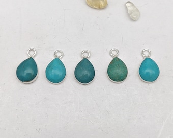 Op de afbeelding: Vijf turquoise druppelvormige hangers met zilveren lussen. De hangers zijn in een rij op een wit oppervlak gerangschikt.