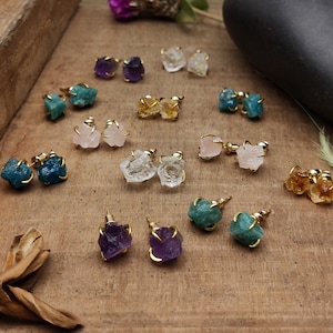 May include: A collection of gemstone stud earrings in various colors, including amethyst, clear quartz, rose quartz, and citrine. Each earring features a raw, uncut stone set in a gold-colored metal setting. The earrings are displayed on a wooden surface.