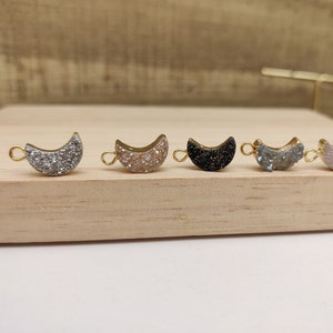 May include: Five gold-toned crescent moon charms with different colored glitter finishes. The charms are arranged in a row on a wooden surface. The colors are silver, gold, black, blue, and white.