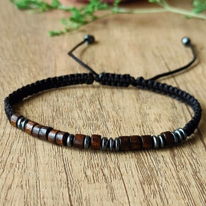 Puede incluir: Un brazalete de cordón negro con cuentas marrones y negras. El brazalete es ajustable y tiene un nudo deslizante.