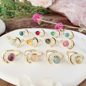 May include: A set of 10 gold crescent moon rings with different colored gemstones. The rings are arranged on a white plate with a pink and white flower in the foreground.