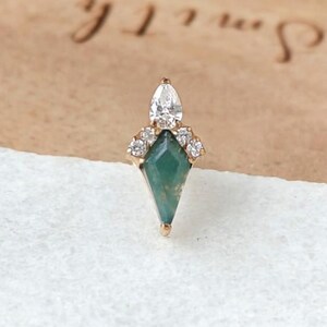 May include: A gold-toned earring with a geometric design. The earring features a teardrop-shaped clear stone at the top, a cluster of small clear stones, and a green, diamond-shaped stone at the base. The earring is set against a light-colored background.
