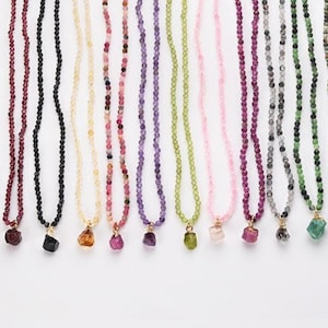 Puede incluir: Una colección de collares de cuentas en varios colores, incluyendo rojo, negro, amarillo, rosa, morado, verde y azul. Cada collar presenta una cadena dorada y un colgante de piedra preciosa. Los collares se exhiben sobre un fondo blanco.