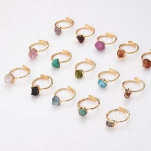 May include: A collection of adjustable gold-tone rings, each featuring a raw, uncut gemstone. The gemstones vary in color, including pink, green, blue, and brown. The rings are displayed on a white surface.