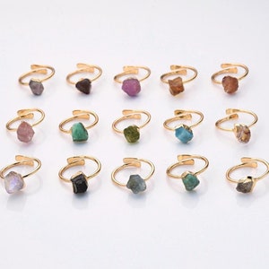 May include: An assortment of gold-coloured adjustable rings, each with a distinctive, irregularly shaped gemstone. The gemstones are in various colours, such as purple, green, brown, and blue. The rings are presented on a white surface.