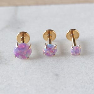 May include: Three gold-colored stud earrings with iridescent, light purple gemstones. The earrings are displayed on a white marble surface. The gemstones have a round shape and vary in size.
