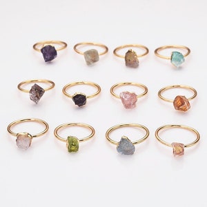 May include: Twelve gold rings each featuring a different coloured raw, uncut gemstone. The gemstones include amethyst, aquamarine, citrine, emerald, garnet, peridot, rose quartz and smoky quartz.