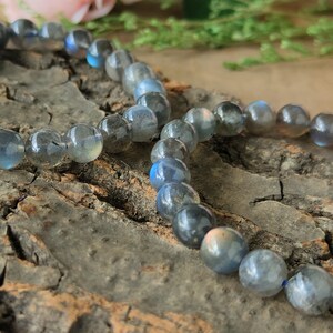 May include: A beaded bracelet made of iridescent blue labradorite stones. The bracelet is laid out on a piece of tree bark.