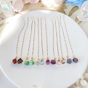 Puede incluir: Una colección de pendientes de hilo dorados, cada uno adornado con una piedra preciosa en bruto de diferente color. Las piedras preciosas incluyen tonos de rojo, verde, morado y azul. Los pendientes se exhiben sobre una superficie blanca.