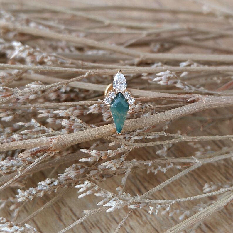 May include: A gold-toned earring with a geometric design. The earring features a teal-colored, diamond-shaped stone at the base, topped with a cluster of clear stones and a larger, oval-shaped clear stone. The earring is set against a backdrop of dried, light brown twigs.