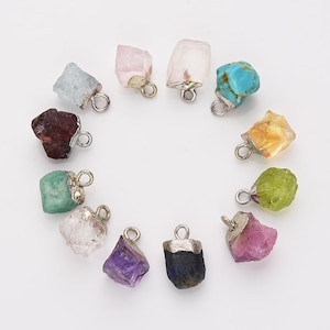 May include: Assortment of 12 raw gemstone pendants with silver jump rings. The gemstones include amethyst, aquamarine, citrine, emerald, garnet, peridot, pink tourmaline, rose quartz, ruby, sapphire, and turquoise.