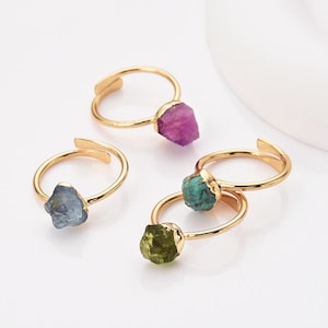 May include: Four gold-colored adjustable rings, each featuring a different rough-cut gemstone. The gemstones are various colors, including purple, blue, green, and turquoise. The rings are displayed on a white surface.