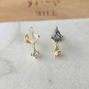 以下が含まれることがあります： ゴールドカラーのスタッドピアス。各イヤリングは、ダイヤモンド型の宝石を特徴とし、1つはクリア、もう1つは緑色のインクルージョンがあり、小さな、ぶら下がっている、正方形にカットされた、クリアな宝石から吊り下げられています。白い表面にあります。
