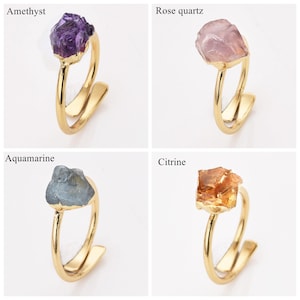 May include: Four gold-colored rings, each featuring a different gemstone: amethyst, rose quartz, aquamarine, and citrine. The gemstones are rough-cut and mounted on a minimalist, open-style band.