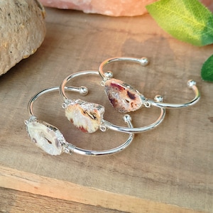 May include: Three silver-toned cuff bracelets, each featuring a raw, irregular agate stone slice at the center. The stones display varied earthy tones of white, brown, and orange with crystalline textures. The metal bands have ball-end terminals.