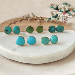 May include: A collection of gemstone stud earrings in various shapes and colors. The earrings feature turquoise and green stones set in gold-colored metal. The earrings are displayed on a white and wood-toned surface, with floral accents in the background.