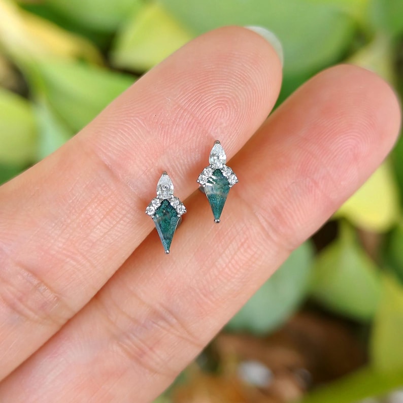 May include: A pair of diamond-shaped earrings with a teal-green center stone and a white diamond accent. The earrings are set in a silver metal. The earrings are displayed on a finger against a blurred green background.