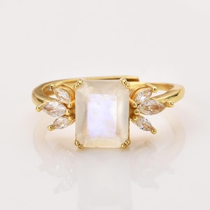 May include: A gold-toned ring featuring a central rectangular moonstone with an iridescent sheen. The stone is flanked on both sides by three small, clear marquise-cut stones arranged in a fan-like pattern. The band is thin and metallic.
