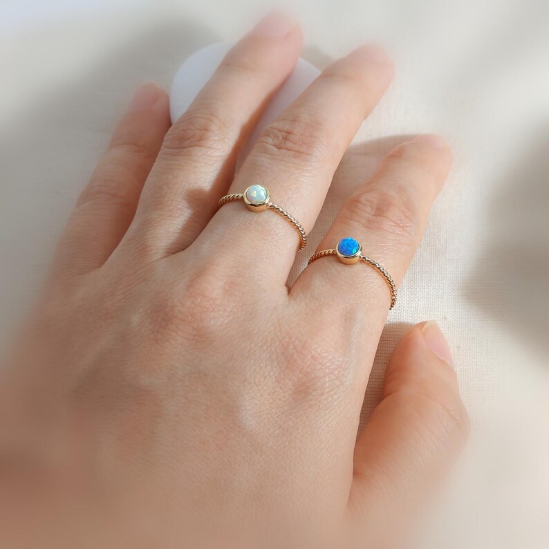 Puede incluir: Dos anillos de oro con bandas trenzadas, cada uno con una piedra redonda engastada. Un anillo muestra un &oacute;palo blanco lechoso, mientras que el otro presenta un &oacute;palo azul vibrante. Los anillos se llevan en dedos adyacentes, resaltando la textura.