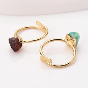 May include: Two adjustable gold rings, one with a rough-cut, dark red gemstone and the other with a light green gemstone. The rings have a simple, elegant design, perfect for everyday wear.