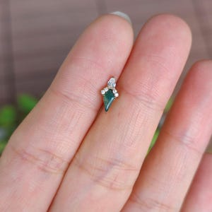 May include: A small, gold-toned earring with a dark green, kite-shaped stone at its center. The earring is adorned with small, clear gemstones. The design is delicate and elegant, suitable for a variety of occasions.