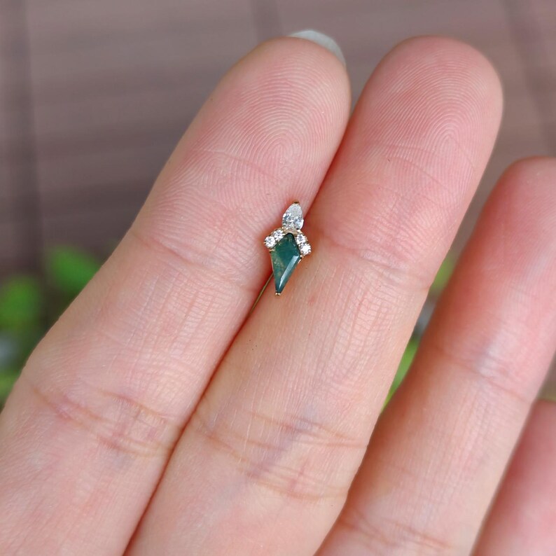 May include: A small, gold-toned earring with a dark green, kite-shaped stone at its center. The earring is adorned with small, clear gemstones. The design is delicate and elegant, suitable for a variety of occasions.