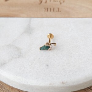 May include: A gold-colored stud earring with a geometric design. The earring features a dark green, triangular stone, a small cluster of clear crystals, and a flat, round backing.