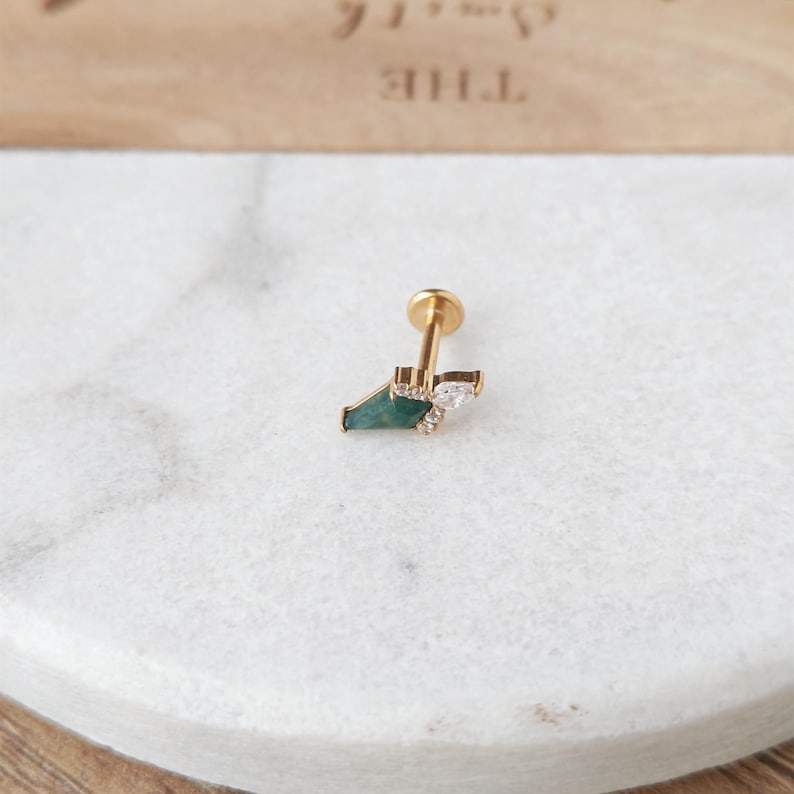 May include: A gold-colored stud earring with a geometric design. The earring features a dark green, triangular stone, a small cluster of clear crystals, and a flat, round backing.