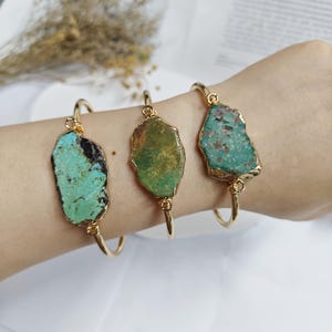 May include: Three gold-toned wire bangles, each featuring a single, irregular, raw-edged stone slab. The stones display varied natural textures and colors, including shades of bright turquoise, earthy olive green, and mottled seafoam green.