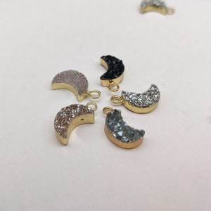 May include: Five crescent moon shaped charms with gold jump rings. The charms are made of different colours of agate, including black, white, grey, and pink.