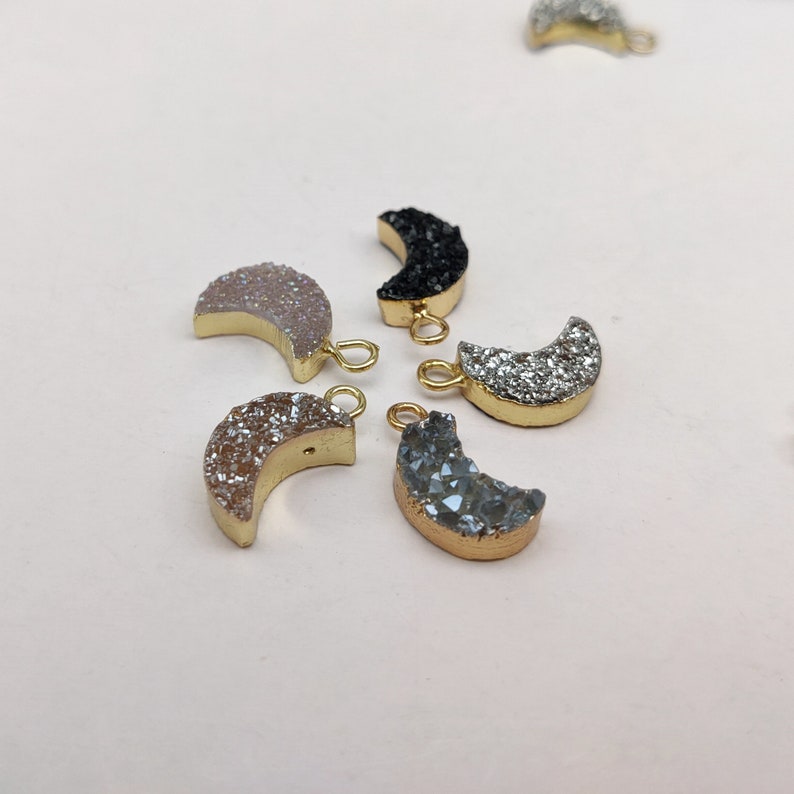May include: Five crescent moon shaped charms with gold jump rings. The charms are made of different colours of agate, including black, white, grey, and pink.