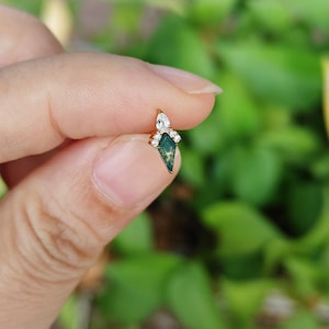 May include: A gold-colored earring with a green, diamond-shaped stone and small, clear accent stones. The earring has a unique design and is held between fingers against a blurred green background.