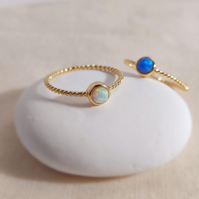 Puede incluir: Dos anillos de oro con bandas trenzadas y piedras engastadas. El anillo delantero presenta una piedra redonda blanca iridiscente, mientras que el anillo situado detr&aacute;s muestra una piedra redonda de color azul vibrante, ambos en metal dorado.