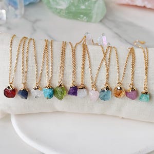 Op de afbeelding: Een verzameling van 12 gouden kettingen met ruwe kristallen hangers. De kristallen zijn in verschillende kleuren, waaronder rood, blauw, wit, turquoise, groen, paars, roze en lichtblauw.