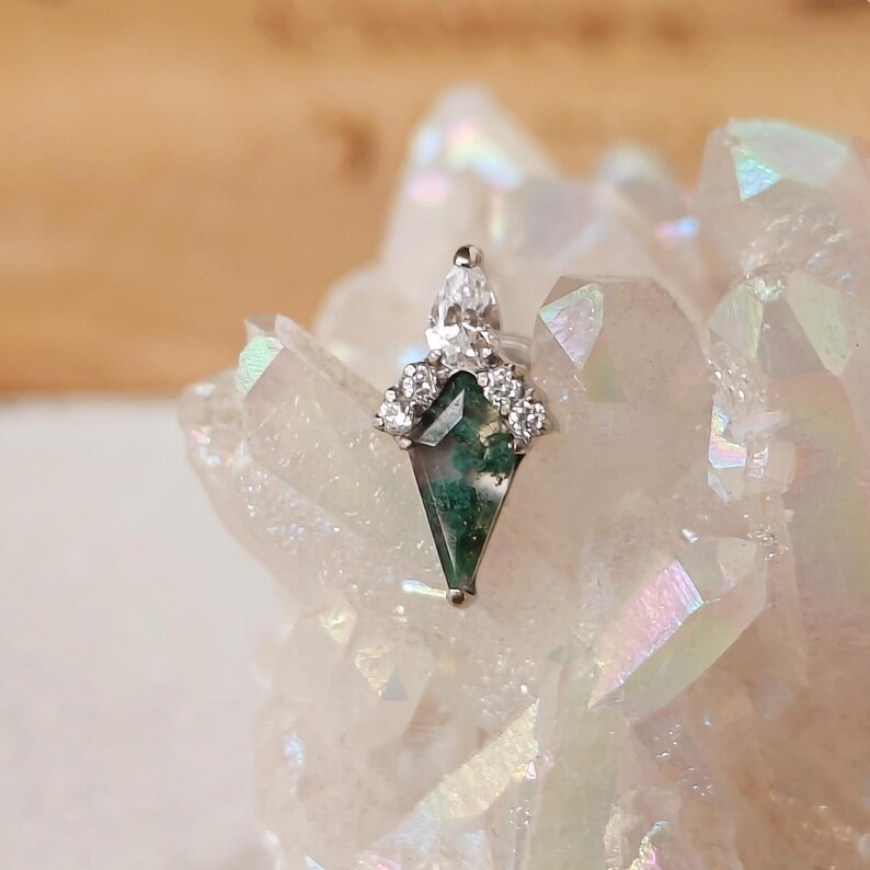May include: A silver-toned earring with a geometric, emerald-green stone and clear accent stones. The earring has a unique shape and is set against a backdrop of a crystal formation.