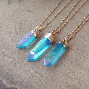 May include: Three gold-toned necklaces with iridescent blue crystal pendants. The crystals have a pointed shape and are capped with gold. The chains are delicate and the pendants are arranged on a wooden surface.