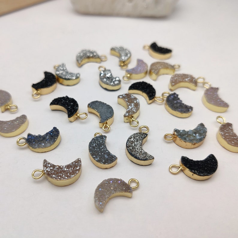 May include: A collection of 20 gold-plated crescent moon charms made of agate. The charms are in various shades of grey, black, and white with a sparkly, glittery finish.