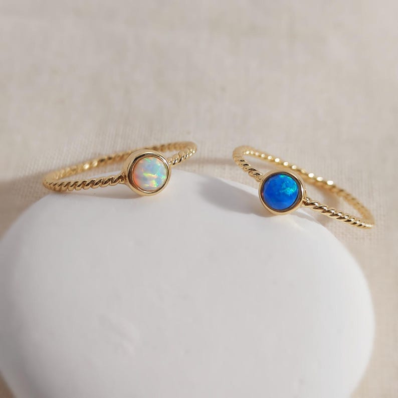 Puede incluir: Dos anillos dorados con dise&ntilde;o de banda trenzada. Un anillo presenta una piedra de &oacute;palo blanca iridiscente redonda, mientras que el otro anillo presenta una piedra redonda de color azul vibrante. Ambas piedras est&aacute;n engastadas en oro.