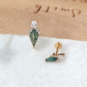 Könnte beinhalten: Zwei goldfarbene Ohrringe. Jeder Ohrring hat einen grünen, rautenförmigen Stein mit weißen Akzenten und einen klaren, tropfenförmigen Stein. Die Ohrringe sind auf einer weißen Oberfläche und einem hellbraunen Hintergrund platziert.