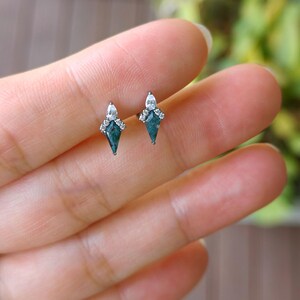 May include: A pair of small, silver-toned stud earrings. Each earring features a dark teal, kite-shaped stone, accented with a cluster of small, clear crystals at the top. The earrings are displayed against a blurred background.