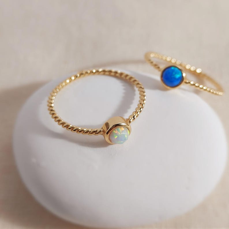 Puede incluir: Dos anillos dorados con bandas trenzadas. Un anillo presenta una piedra redonda de color blanco iridiscente, mientras que el otro anillo tiene una piedra redonda de color azul vibrante. Ambos anillos tienen un acabado pulido y un dise&ntilde;o sencillo.