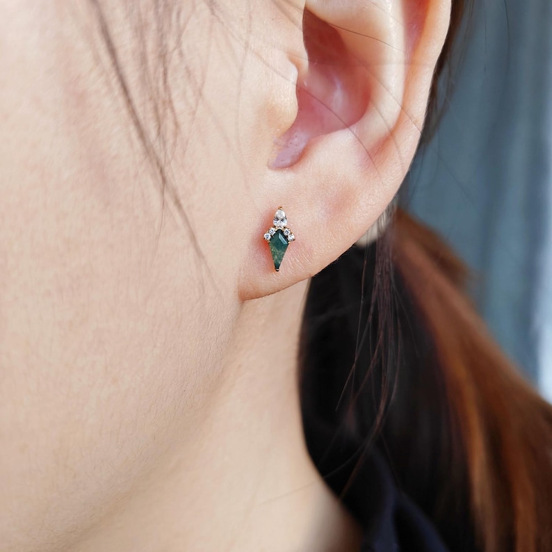 May include: A gold stud earring featuring a green gemstone in a kite shape, accented with small white gemstones. The earring is worn on an ear, showcasing its design and color contrast. The earring's design is elegant and eye-catching.
