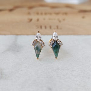 May include: A pair of gold stud earrings. Each earring features a teardrop-shaped green gemstone with a white pear-shaped stone at the top, surrounded by small clear stones. The earrings are set on a white surface.
