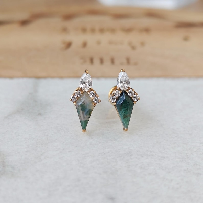 May include: A pair of gold stud earrings. Each earring features a teardrop-shaped green gemstone with a white pear-shaped stone at the top, surrounded by small clear stones. The earrings are set on a white surface.