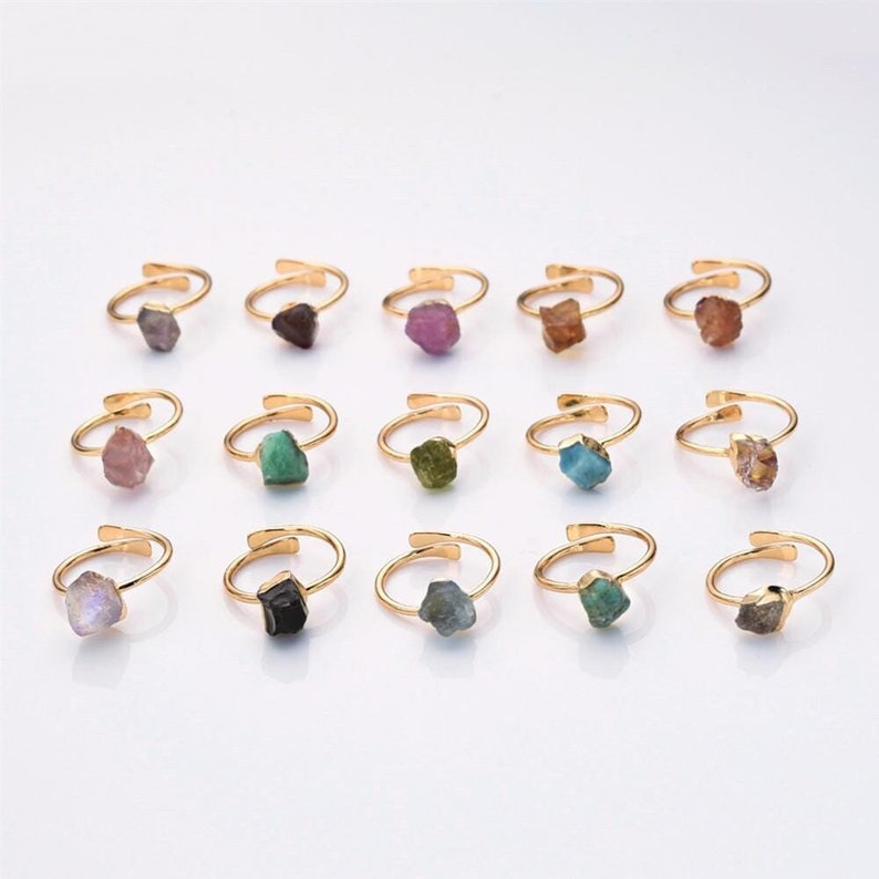 May include: A collection of adjustable gold-tone rings, each featuring a unique, irregularly shaped gemstone. The gemstones vary in color, including purple, green, brown, and blue. The rings are displayed on a white surface.